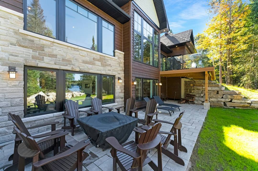 Large paver patio with Adirondack chairs around a fire table beside a stone-veneer home